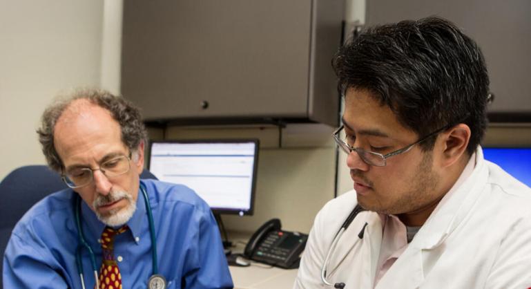 Two physicians reviewing information together in a medical office, with computers and phones in the background.