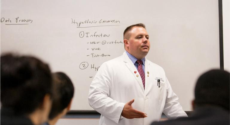 Physician in a white coat giving a lecture to medical students, standing in front of a whiteboard with handwritten notes.