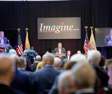 Photo taken from back of room, back of heads blurred in foreground. Speaker is shown under Project Imagine logo on overhead screen.