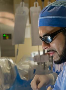 Surgeon in scrubs, cap, and protective glasses focused during a medical procedure.